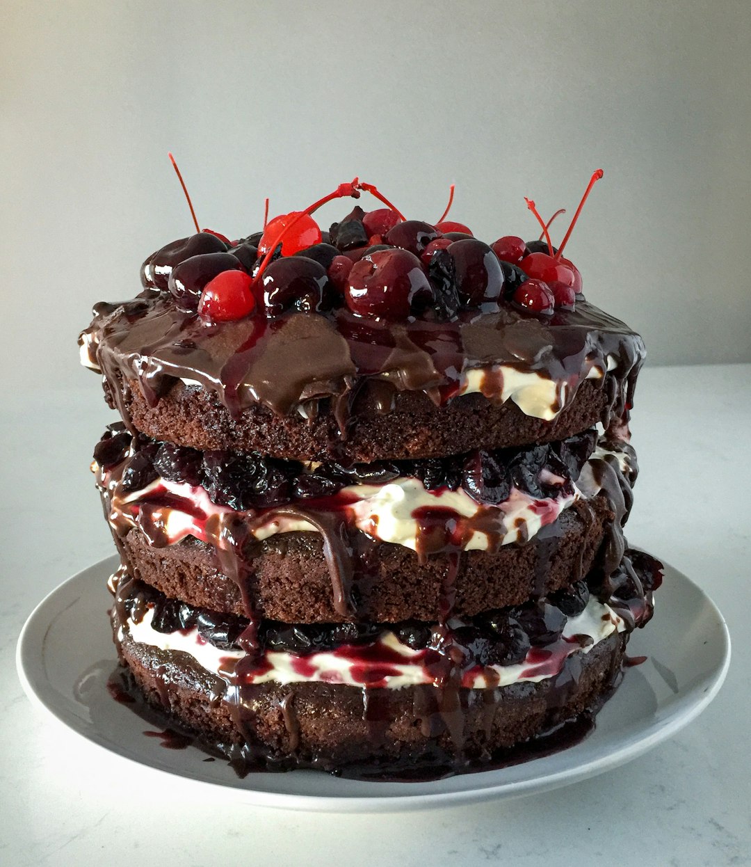 A three-layer chocolate cake with cream, dark cherries, and red glaze on a plate